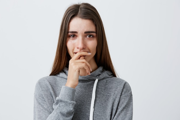 Emociones negativas. Retrato de joven encantadora bonita chica caucásica con cabello largo castaño en moda casual con capucha llorando, boca de ropa con la mano, sentirse deprimido y aplastado