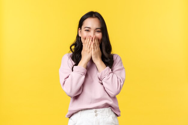 Las emociones de las personas, el estilo de vida y el concepto de moda. Mujer coreana divertida y linda riendo tímida, sonriendo con los ojos mientras se tapa la boca y ríe tontamente a la cámara, de pie con fondo amarillo.