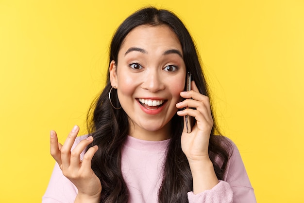 Las emociones de las personas, el estilo de vida, el ocio y el concepto de belleza. Alegre niña asiática recibe noticias maravillosas por teléfono, hablando por teléfono móvil y sonriendo asombrada, levanta la mano sorprendida y feliz, fondo amarillo.