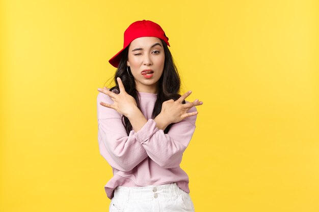 Las emociones de las personas, el estilo de vida, el ocio y el concepto de belleza. Chica asiática con estilo fresco y atrevido en gorra roja de hip hop, mostrando gesto de botín y sonrisa atrevida, de pie confiado sobre fondo amarillo.