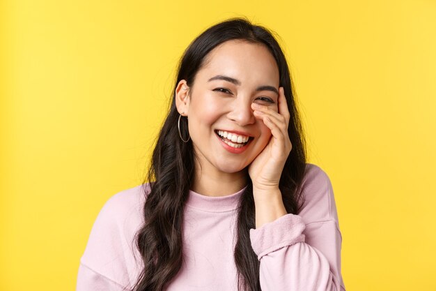 Las emociones de las personas, el estilo de vida, el ocio y el concepto de belleza. Hermosa chica asiática toca la cara y ríe con una sonrisa alegre y feliz, escucha cumplidos y sonrojo, soporte fondo amarillo.