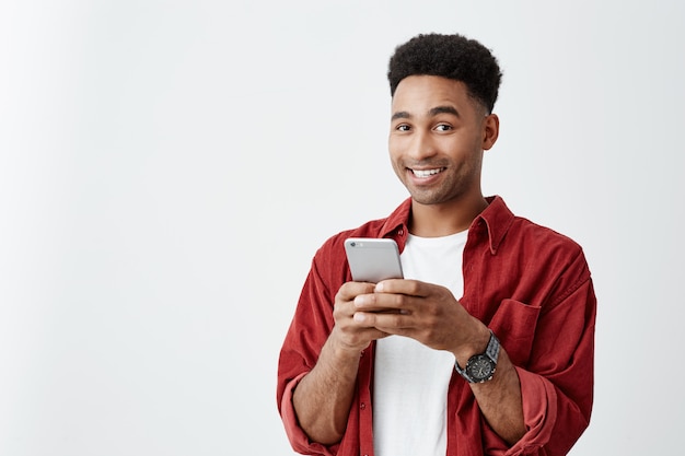 Emociones positivas. Cerca de joven apuesto hombre de piel oscura con peinado afro en camiseta blanca y camisa roja sonriendo con dientes, conversando con un amigo en el teléfono inteligente