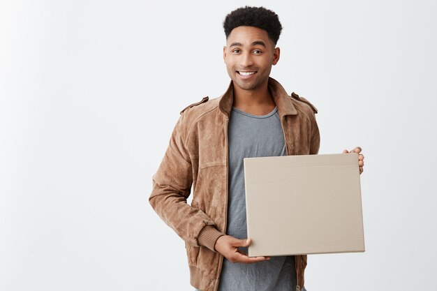 Emociones positivas. Cerca de joven hermoso hombre de piel negra con peinado afro en ropa casual elegante con cartón en las manos, mirando a la cámara con expresión feliz y relajada.