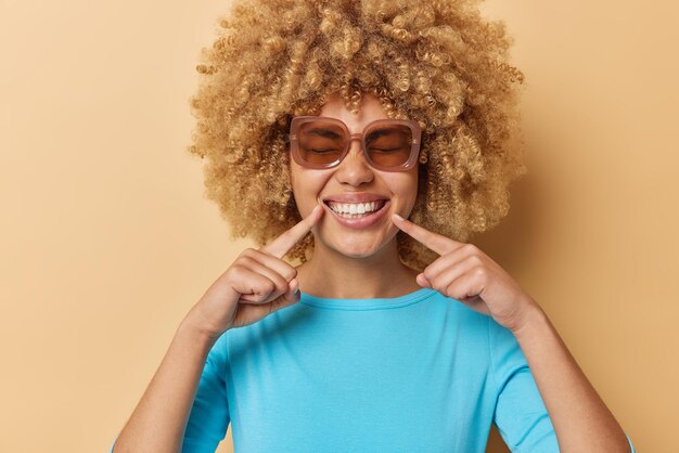 Emociones positivas y concepto de expresiones faciales La mujer de pelo rizado feliz señala una sonrisa brillante con dientes que muestra dientes blancos perfectos, usa gafas de sol y un puente azul aislado sobre fondo marrón