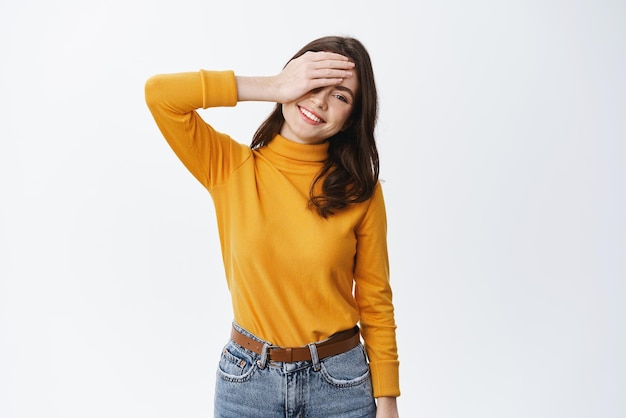 Emociones positivas Mujer joven feliz y despreocupada sosteniendo la mano en la cara sonriendo y mirando alegre a la cámara de pie sobre fondo blanco.