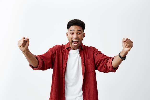 Emociones positivas. Retrato de un hombre joven y feliz de piel oscura con corte de pelo afro en traje casual elegante extendiendo las manos con entusiasmo, gritando, animando al equipo de fútbol favorito.