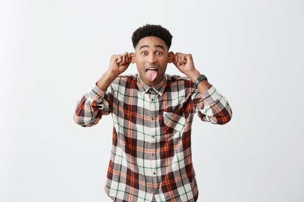 Emociones positivas. Retrato de joven atractivo hombre de piel oscura con peinado afro en camisa casual a cuadros con orejas, mostrando la lengua, haciendo muecas para hacer reír al niño pequeño en la calle.