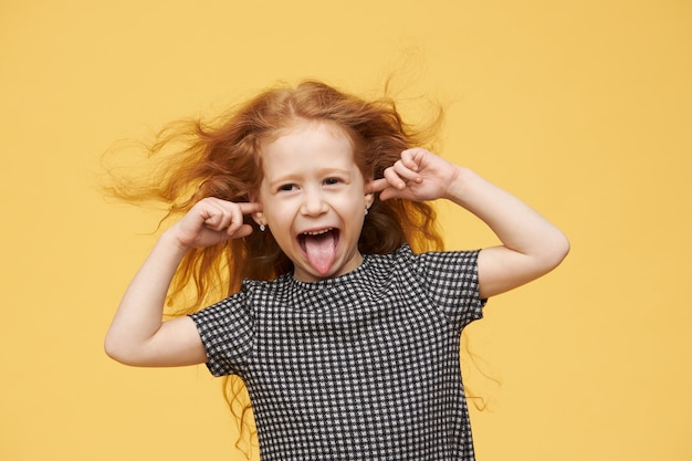 Emociones, reacciones y lenguaje corporal genuinos del ser humano. Niña mimada enojada con cabello rojo sacando la lengua, fingiendo no escucharte, tapando los oídos, gritando, enojada y traviesa