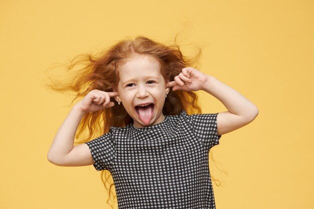 Emociones, reacciones y lenguaje corporal genuinos del ser humano. Niña mimada enojada con cabello rojo sacando la lengua, fingiendo no escucharte, tapando los oídos, gritando, enojada y traviesa