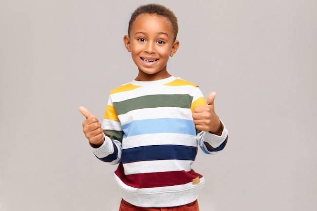 Emociones, reacciones y sentimientos humanos positivos. Niño de piel oscura feliz emocional en jersey multicolor haciendo gesto de pulgar hacia arriba, expresando su acuerdo, aprobación, dando su gusto, sonriendo ampliamente