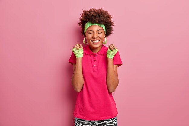 Emociones y sentimientos felices. Sonriente niña afroamericana con camiseta rosa, guantes deportivos y diadema, aprieta los puños con alegría, siente el sabor de la victoria, celebra el concurso ganador