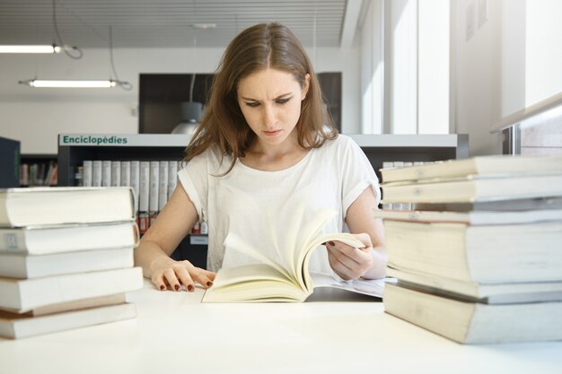 Emociones y sentimientos humanos. Mujer estudiante estresada preparándose para los exámenes finales, estudiando en la biblioteca frente a grandes montones de libros, mirando el libro de texto