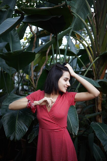 Emociones y sentimientos humanos. Retrato de mujer joven hermosa positiva con elegante vestido rojo, disfrutando de la frescura proveniente de las plantas verdes, sonriendo con alegría, sintiéndose inspirado por la naturaleza