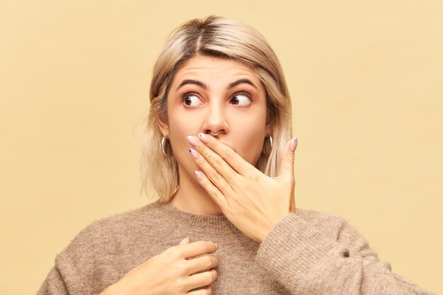 Emociones, sentimientos y reacciones humanas genuinas. Atractiva joven rubia con mirada de sorpresa, tratando de mantener información secreta o confidencial, cubriendo la boca con la mano y mirando a otro lado