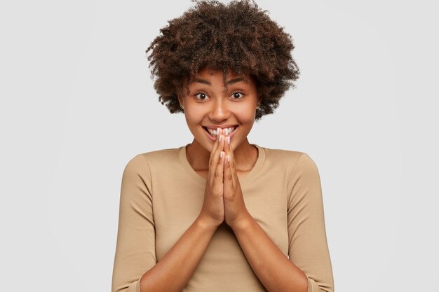 Emotiva y complacida niña de piel oscura tiene una sonrisa con dientes, mantiene las manos en gesto de oración