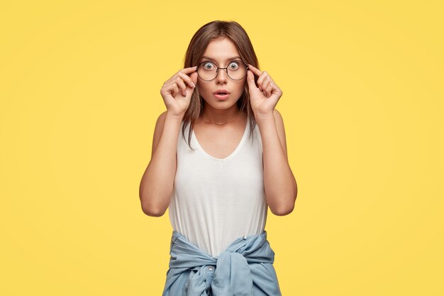 Emotiva morena joven horrorizada con gafas posando contra la pared amarilla