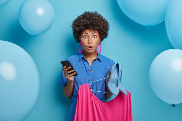 Emotiva mujer asustada con ropa elegante, elige vestido y zapatos de tacón para vestirse en la fiesta de cumpleaños, sostiene el teléfono inteligente