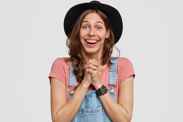 Emotiva mujer europea en ropa casual, mantiene las manos juntas sobre el pecho, sonríe con alegría al escuchar las buenas noticias, usa sombrero negro y peto, modelos contra la pared blanca