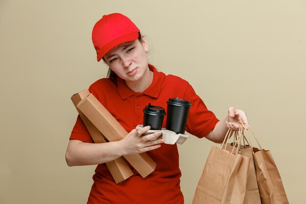 Empleada de entrega con gorra roja y uniforme de camiseta en blanco con bolsas de papel y tazas de café que parecen confundidas y disgustadas de pie sobre fondo marrón
