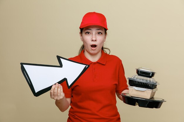 Empleada de entrega con gorra roja y uniforme de camiseta en blanco sosteniendo contenedores de comida y una gran flecha mirando a la cámara asombrada y sorprendida de pie sobre fondo marrón
