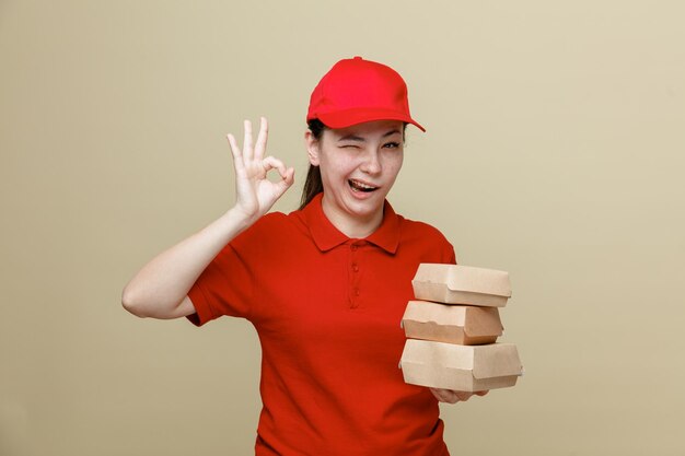 Empleada de entrega con gorra roja y uniforme de camiseta en blanco sosteniendo contenedores de comida mirando a la cámara feliz y positiva sonriendo alegremente mostrando el signo de ok guiñando un ojo de pie sobre fondo marrón