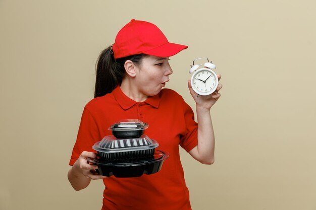 Empleada de entrega con gorra roja y uniforme de camiseta en blanco sosteniendo recipientes de comida y despertador mirándolo asombrado y sorprendido de pie sobre fondo marrón