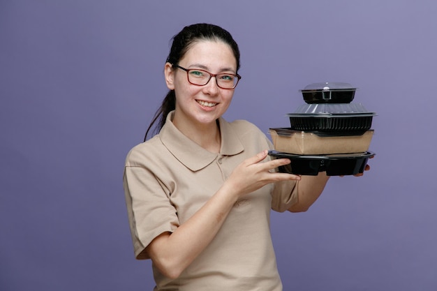 Empleada de entrega en uniforme de polo en blanco con gafas sosteniendo contenedores de comida mirando a la cámara feliz y positiva sonriendo alegremente de pie sobre fondo azul