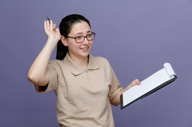 Empleada de entrega en uniforme de polo en blanco con gafas sosteniendo portapapeles mirando a un lado feliz y positiva sonriendo alegremente de pie sobre fondo azul