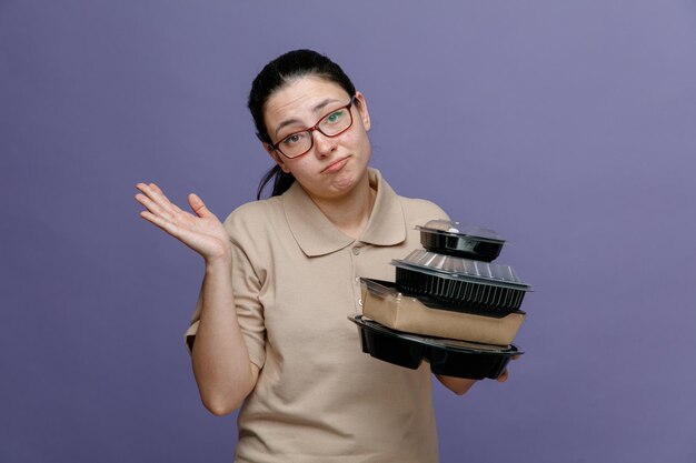 Empleada de entrega en uniforme de polo en blanco con gafas sosteniendo recipientes de comida que parece confundida con dudas levantando el brazo de pie sobre fondo azul