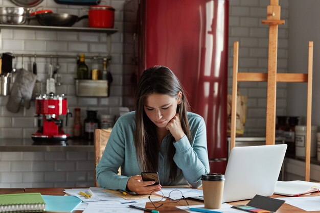 Empleada femenina con cabello largo oscuro, vestida con una camisa elegante, usa un teléfono móvil, trabaja en un informe comercial, bebe café para llevar, usa una computadora portátil,