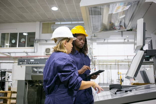 Empleada industrial femenina enseñando a su colega a operar la máquina, apuntando al tablero de control, usando tableta