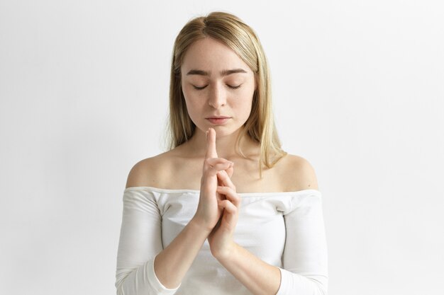 Empleada joven atractiva meditando en la oficina blanca, manteniendo los ojos cerrados y juntando las manos en gesto, tratando de encontrar el equilibrio dentro de sí misma, practicando ejercicios de respiración