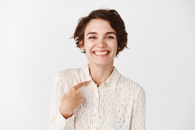 Foto gratuita empleada sonriente que parece feliz mientras se señala a sí misma de pie en una blusa informal sobre fondo blanco copiar espacio