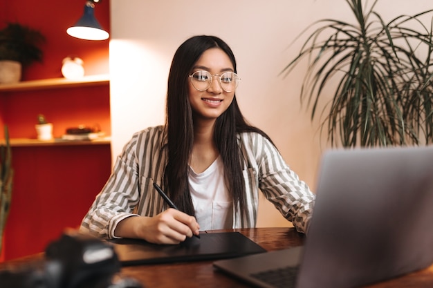 Empleada sonriente en traje de rayas y gafas mira a la computadora portátil y sostiene la pluma