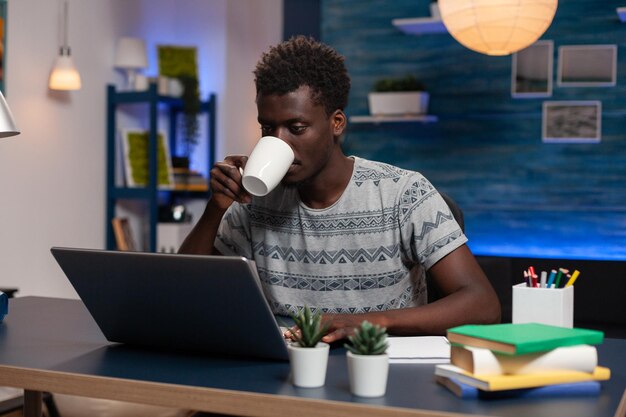 Empleado afroamericano sosteniendo una taza de café escribiendo ideas de marketing en la computadora trabajando remotamente en un proyecto en línea en la sala de estar. Freelancer sentado en el escritorio analizando gráfico financiero