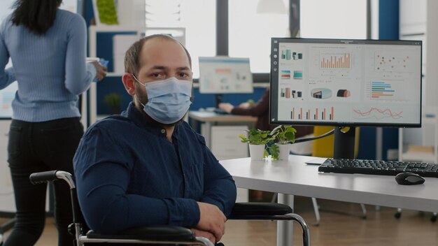 Empleado con discapacidades, inválido, discapacitado paralizado con mascarilla contra el coronavirus mirando a la cámara sentado inmovilizado en silla de ruedas en la sala de la oficina de negocios, trabajando en un proyecto financiero