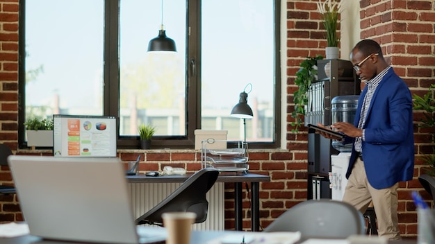 Foto gratuita empleado de la empresa paseando por la oficina de inicio, usando una tableta digital para analizar las estadísticas del proyecto. joven empresario esperando en el espacio de trabajo corporativo para recibir noticias, usando un dispositivo.