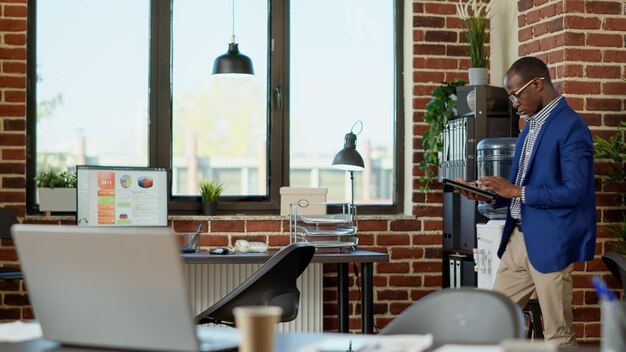 Empleado de la empresa paseando por la oficina de inicio, usando una tableta digital para analizar las estadísticas del proyecto. Joven empresario esperando en el espacio de trabajo corporativo para recibir noticias, usando un dispositivo.