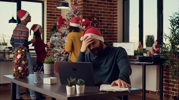 Empleado frustrado por la festividad navideña en la oficina de inicio, tratando de trabajar en una computadora portátil durante la víspera de Navidad. Hombre estresado siendo perturbado e interrumpido por gente ruidosa.