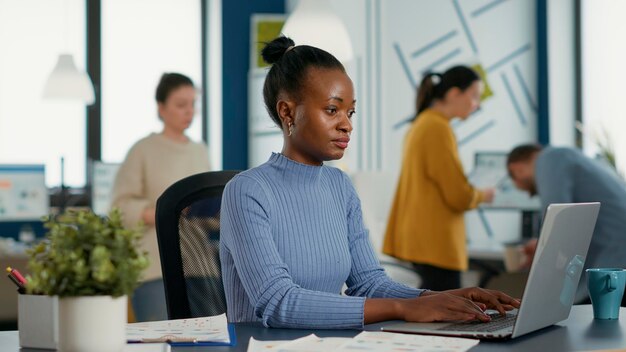 Empleado de inicio afroamericano sentado en el escritorio abriendo una computadora portátil y comenzando a escribir en el teclado trabajando en estadísticas de ventas. Mujer de negocios en una oficina moderna y ocupada que comienza a trabajar por la mañana.