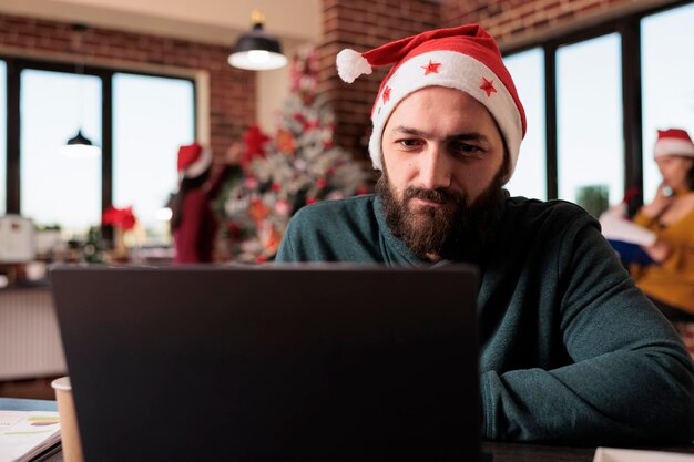 Empleado de inicio que usa una computadora portátil en la oficina comercial para trabajar en un proyecto de la empresa durante la temporada de invierno. Hombre con gorro de Papá Noel trabajando en un informe en el lugar de trabajo con árbol de Navidad y adornos navideños.