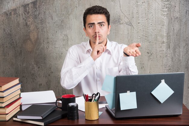 Empleado joven haciendo señal de silencio en el escritorio de oficina.