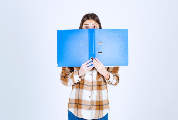 Foto gratuita empleado joven que se esconde detrás de la carpeta azul en la pared blanca.