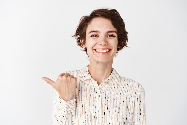 Empleado joven sonriente que parece feliz y orgulloso, señalando con el dedo a la compañía, de pie contra la pared blanca