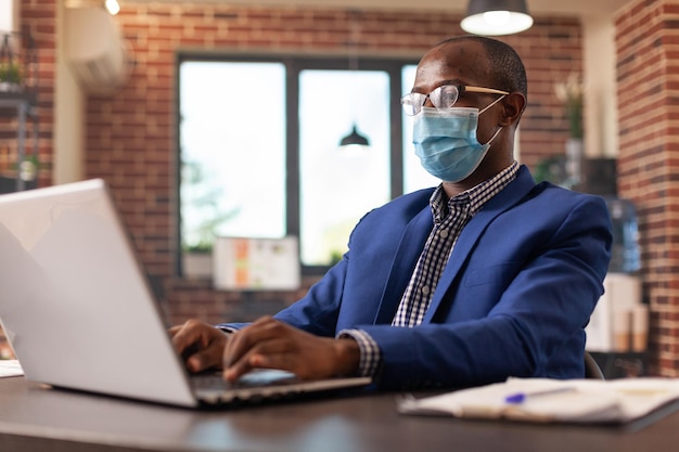 Empleado con mascarilla usando laptop para trabajar en la planificación de proyectos y estrategia comercial. Persona que trabaja en la computadora para hacer trámites y trabajar en una empresa de nueva creación durante la pandemia de covid 19.