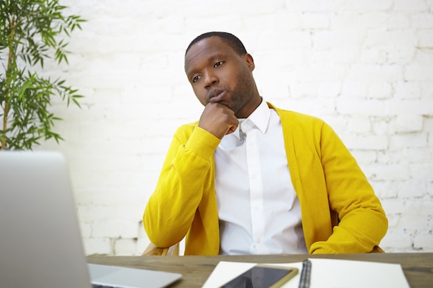 Empleado masculino afroamericano serio vestido elegantemente sentado en la pared de ladrillo blanco en su lugar de trabajo, usando una computadora portátil genérica, frotándose la barbilla, con expresión facial pensativa