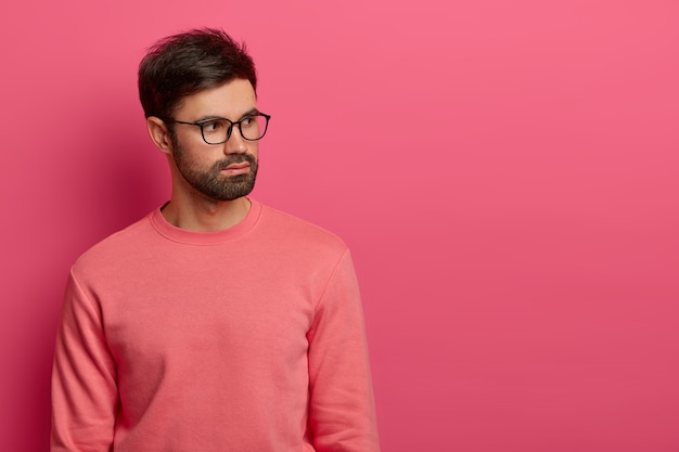 Empleado masculino exitoso, barbudo y de aspecto serio, centrado a un lado del lado derecho, tiene pensamientos profundos sobre problemas laborales futuros, usa lentes transparentes y un jersey rosado, posa en interiores. Disparo monocromo