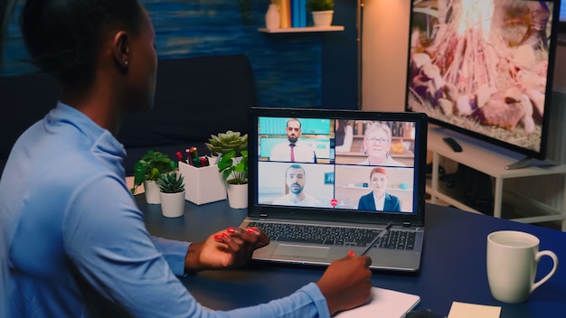 Empleado negro remoto que tiene videoconferencia trabajando desde casa sentado en la sala de estar tarde en la noche. Freelancer que usa la red de tecnología inalámbrica hablando en una reunión virtual a la medianoche haciendo horas extraordinarias