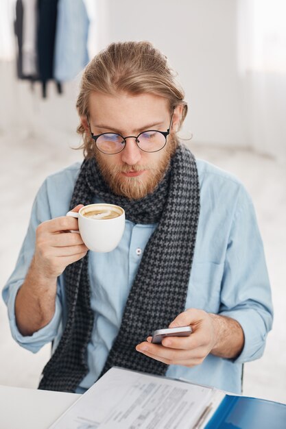 Empleado de oficina masculino barbudo en gafas redondas vestido con camisa azul y bufanda, rodeado de papeles y documentos, recibe el mensaje de negocios en el teléfono inteligente, los tipos responden, bebe café.