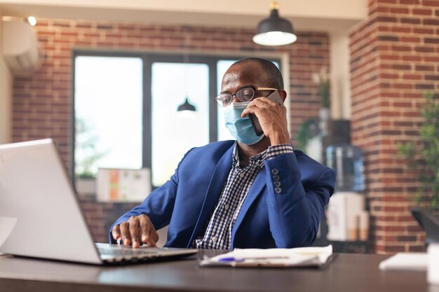 Empleado que usa una llamada telefónica en un teléfono móvil para hablar con el gerente sobre el crecimiento del negocio, usando una mascarilla. Hombre que trabaja con la computadora portátil para planificar el proyecto y hablando por teléfono inteligente en la oficina.